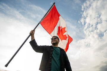 Portrait of cheerful young man looking forward while standing proudly with holding canada flag over his head against wind and blue sky Man holding the canada flag celebration