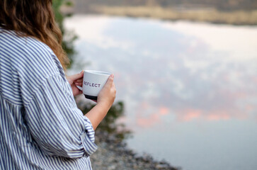 A woman holds a white mug that says “Reflect” by calm water, symbolizing mindfulness,...