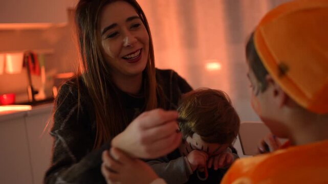 Young mother playfully applying halloween makeup on her two sons, dressed in pumpkin costumes, inside their home