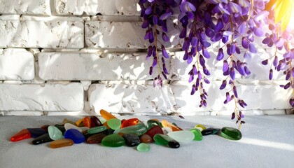 Fototapeta premium A collection of colorful sea glass lies on a surface against a white brick wall, accented by cascading purple wisteria flowers.