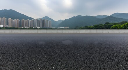 Empty highway asphalt with city and mountain backdrop. A wide, paved road stretches towards a verdant mountain range in the background, with city buildings in the middle ground.