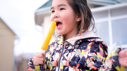 A young girl is about to take a bite of a yellow popsicle. A cute Asian child holds ice cream in...