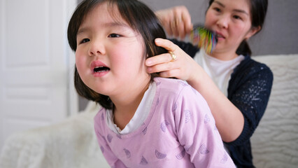 An Asian woman combs the hair of a restless young girl in pink pajamas. Mom makes her little...
