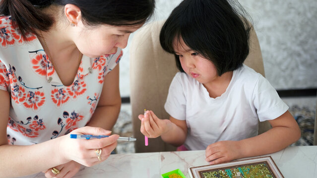A woman helps a little girl with a craft. Mom and daughter are engaged in creative work. They make up a picture of beads