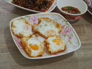 Four crispy fried eggs with runny yolks served on a floral plate, accompanied by spicy chili sauce and stir-fried meat in a vibrant Asian meal setting.