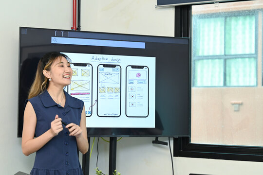 A woman is giving a presentation in front of a large screen. The screen shows a design for a mobile app. Scene is professional and informative