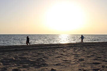 Fototapeta premium People walking along the beach at sunset, silhouetted against the bright sun reflecting on the sea, as the warm golden light illuminates the sandy shoreline. Peaceful coastal evening scene.