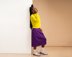 African woman in yellow fuzzy sweater and purple skirt standing indoors, smiling while talking on...