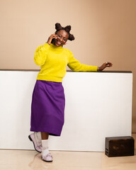 African American woman in yellow sweater and purple skirt smiling while talking on phone indoors,...