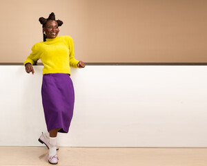 Joyful African woman in yellow sweater and purple skirt leans casually on a white wall in a modern...