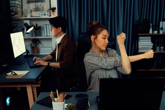 Businesswoman working on desk with stretching arm up and down manner with body health ache of strain overwork while coworker trading stock market on website on pc at late over night time. Infobahn.