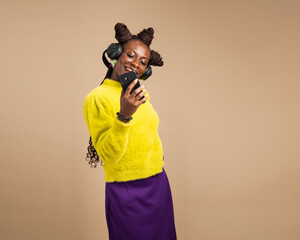 Stylish African woman in yellow sweater with headphones smiles while taking a selfie with smartphone in studio