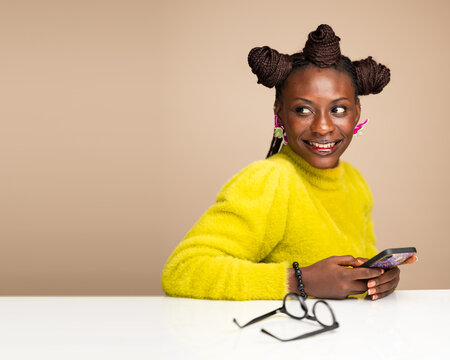 Smiling African American woman in yellow sweater sitting at office desk with smartphone and eyeglasses, looking to the side with joyful expression, workplace technology and lifestyle concept - Powered by Adobe