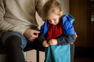 Toddler zipping overall by himself in hallway, father nearby supporting child getting dressed for...