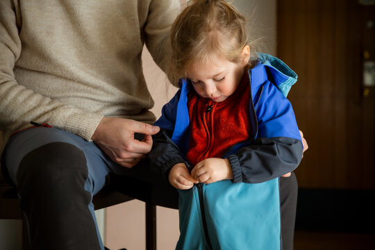 Toddler zipping overall by himself in hallway, father nearby supporting child getting dressed for cold autumn winter outdoor walk.