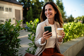 Businesswoman using smartphone and drinking coffee outdoors
