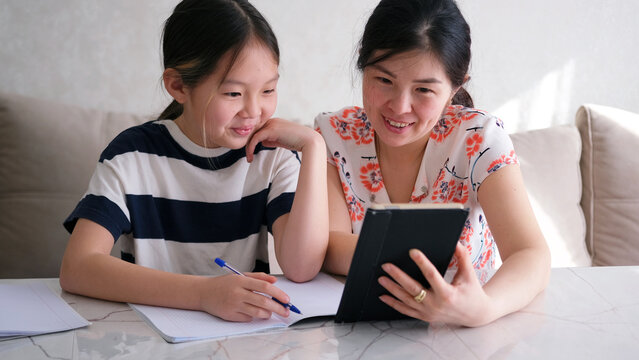 A happy mom and daughter are watching a video math lesson on a tablet. A woman and a girl are sitting at a table and doing their homework - Powered by Adobe
