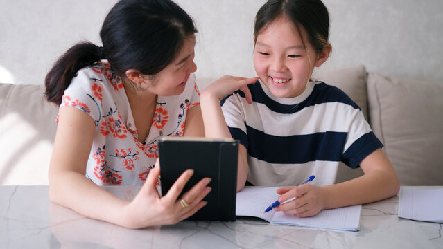 Home education. Tutor. A woman shows a video lesson on her tablet to her little daughter. Happy Asian schoolgirl