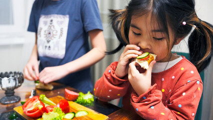 Asian cute girl eats a sandwich with a slice of cucumber and tomato. Her sister is standing behind her. Children snack on homemade sandwiches