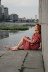 Woman in a burgundy dress sits barefoot by the Moscow River embankment, leaning against a concrete...