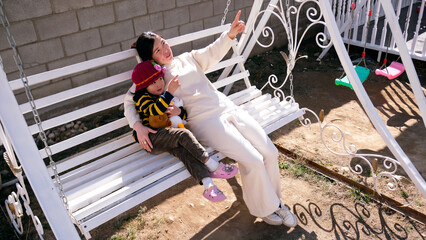 A babysitter sits with a child on a swing. A caring mother talks sweetly with her little daughter. A woman and a baby are swinging on a swing