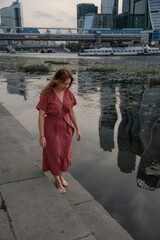 Woman in a burgundy dress walks barefoot along the Moscow River embankment with skyscrapers of...