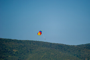A rainbow-colored parasail floats above a lush green hillside under a clear blue sky, capturing a moment of airborne adventure.