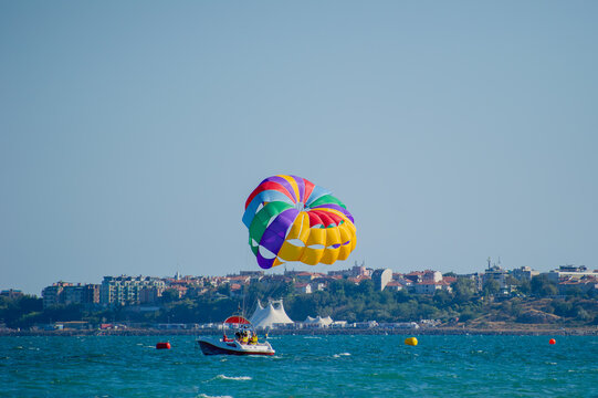 A vibrant parasail glides above the sea, towed by a boat near a lively coastal town under a clear blue sky