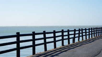 Fototapeta premium A coastal walking path with a black wooden fence casting shadows on the ground, overlooking calm ocean water. Wind turbines are visible on the distant horizon.