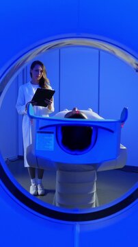 Medical worker standing confidently near MRI tunnel while male patient resting on table. Patient lies on MRI table and undergoes magnetic resonance imaging. Woman in robe and protective cap enters