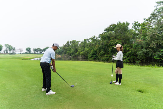 Asian man and woman golfers swing clubs together on scenic golf course.
