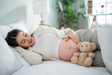 Asian young pregnant woman gently stroking belly while lying down on bed. 