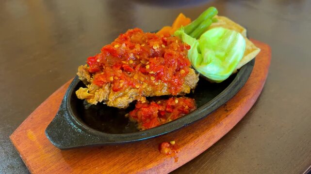 Ayam Geprek or Spicy fried chicken with sambal, served on a hot plate with tofu and fresh vegetables on a wooden table