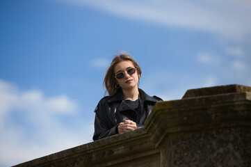 Fototapeta premium pretty young blonde woman with sunglasses, black leather jacket, boots and short skirt leaning against the wall with cool attitude. The woman is smiling and happy. In the background the blue sky.