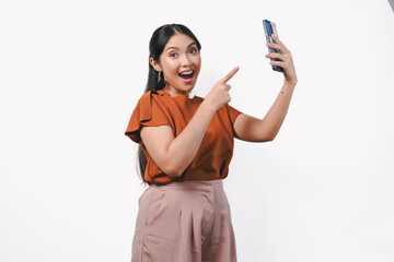 Naklejka premium Joyful young Asian woman in brown shirt pointing to the cellphone she holds with a happy smile.