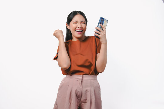Excited young Asian woman raising her fists with happy successful expression while holding phone.