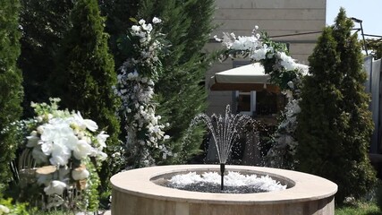 This video captures a beautiful outdoor wedding or event setting, featuring a tiered fountain with water jets amidst lush greenery and elegant floral arrangements. - Powered by Adobe