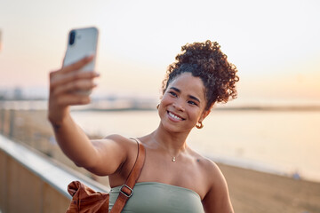 Young tourist capturing a joyful selfie with her smartphone at sunset, enjoying the beautiful beach and the golden hour glow