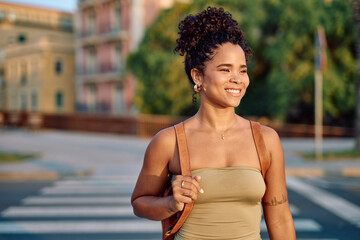 Happy woman with curly hair and brown leather backpack is walking in the city during a sunny summer...