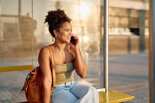 Happy young woman chatting on a mobile phone while sitting at a bus stop on a sunny summer day, enjoying the outdoors and urban vibes