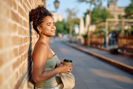 Tourist enjoying a relaxing moment with closed eyes, holding a takeaway coffee and leaning against a brick wall in a city street - Powered by Adobe