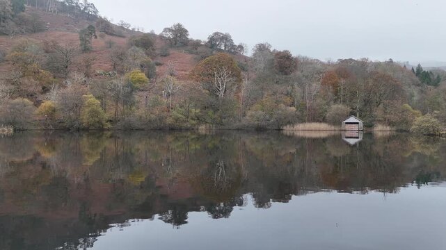 Rydal Water, A serene lake reflects the surrounding trees and a boathouse under an overcast sky, creating a peaceful autumn landscape.