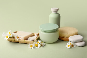 Composition with massage body brush, chamomile flowers, jar and bottle of cosmetic products on green background, closeup