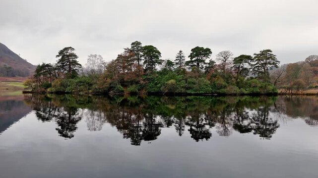Rydal Water England. A tranquil scene of a tree-covered island reflected in a calm lake, with a backdrop of hills under a cloudy sky.