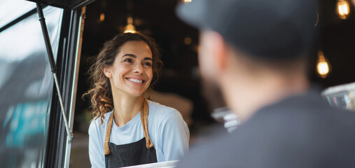 Smiling female vendor in apron talking to customer at food truck window, cheerful interaction highlighting street food culture, service and small business atmosphere. Selective focus.