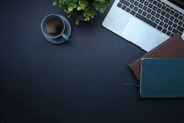 Top view of laptop, coffee cup, notebooks, and green plant on dark desk