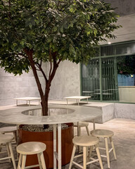 Modern cafe interior with a tree centerpiece, round table, and wooden stools, combining natural and minimalist industrial design.