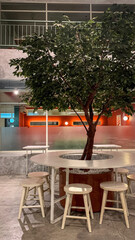 Modern cafe interior with a tree centerpiece, round table, and wooden stools, combining natural and minimalist industrial design.