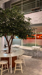 Modern cafe interior with a tree centerpiece, round table, and wooden stools, combining natural and minimalist industrial design.