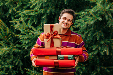 Smiling man in cozy sweater holding wrapped gifts by Christmas tree